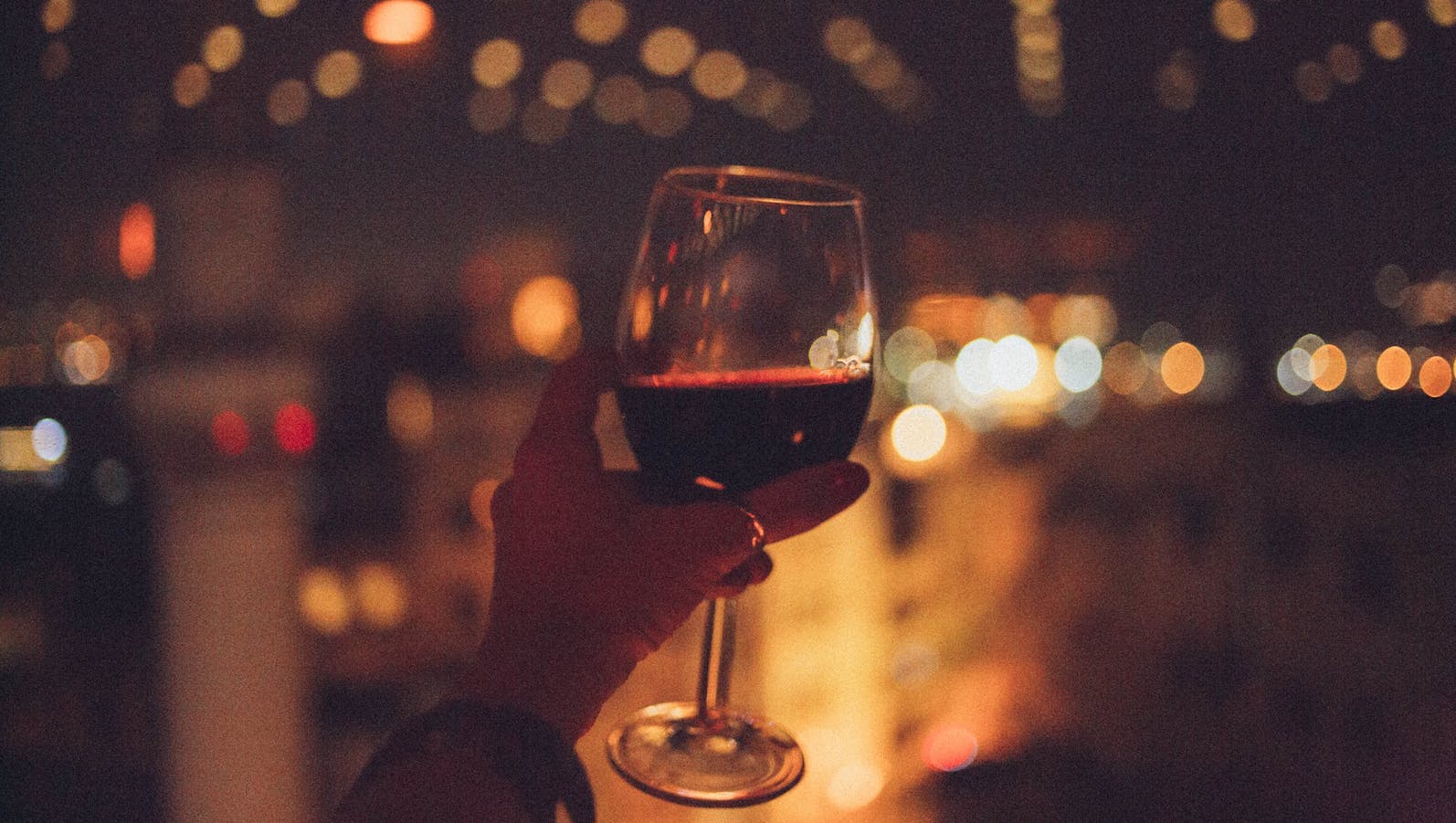 A person's hand holding a glass of red wine in a dark room with some lights