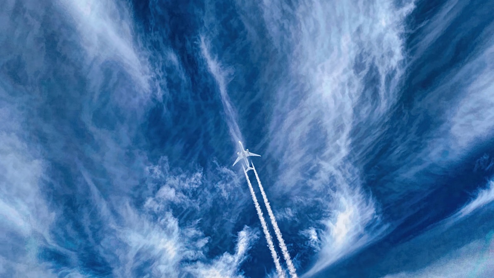 Below view of a plane with a trail against a blue sky with wispy clouds