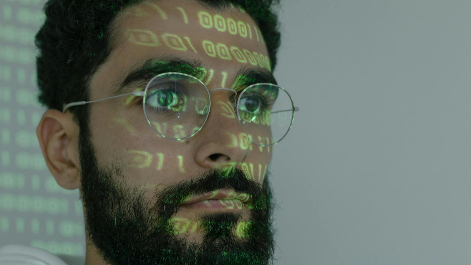 Man in White Crew Neck Shirt Wearing Silver Framed Eyeglasses With Binary Digits Projected on His Face