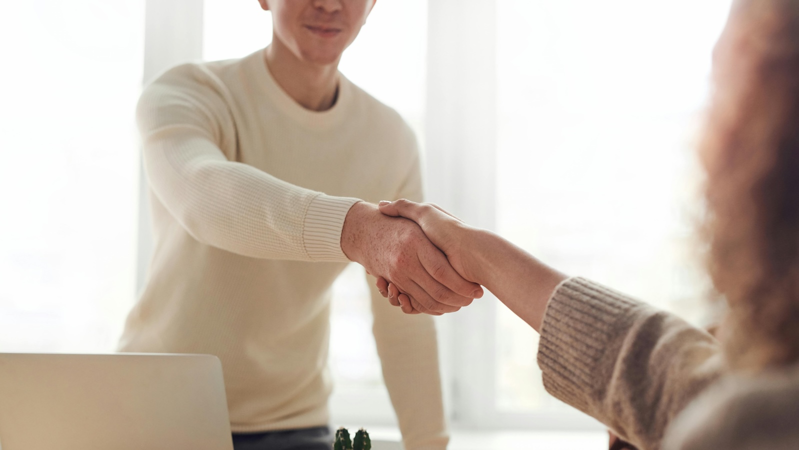 Two people shaking hands over a desk in front of window letting a lot of light in