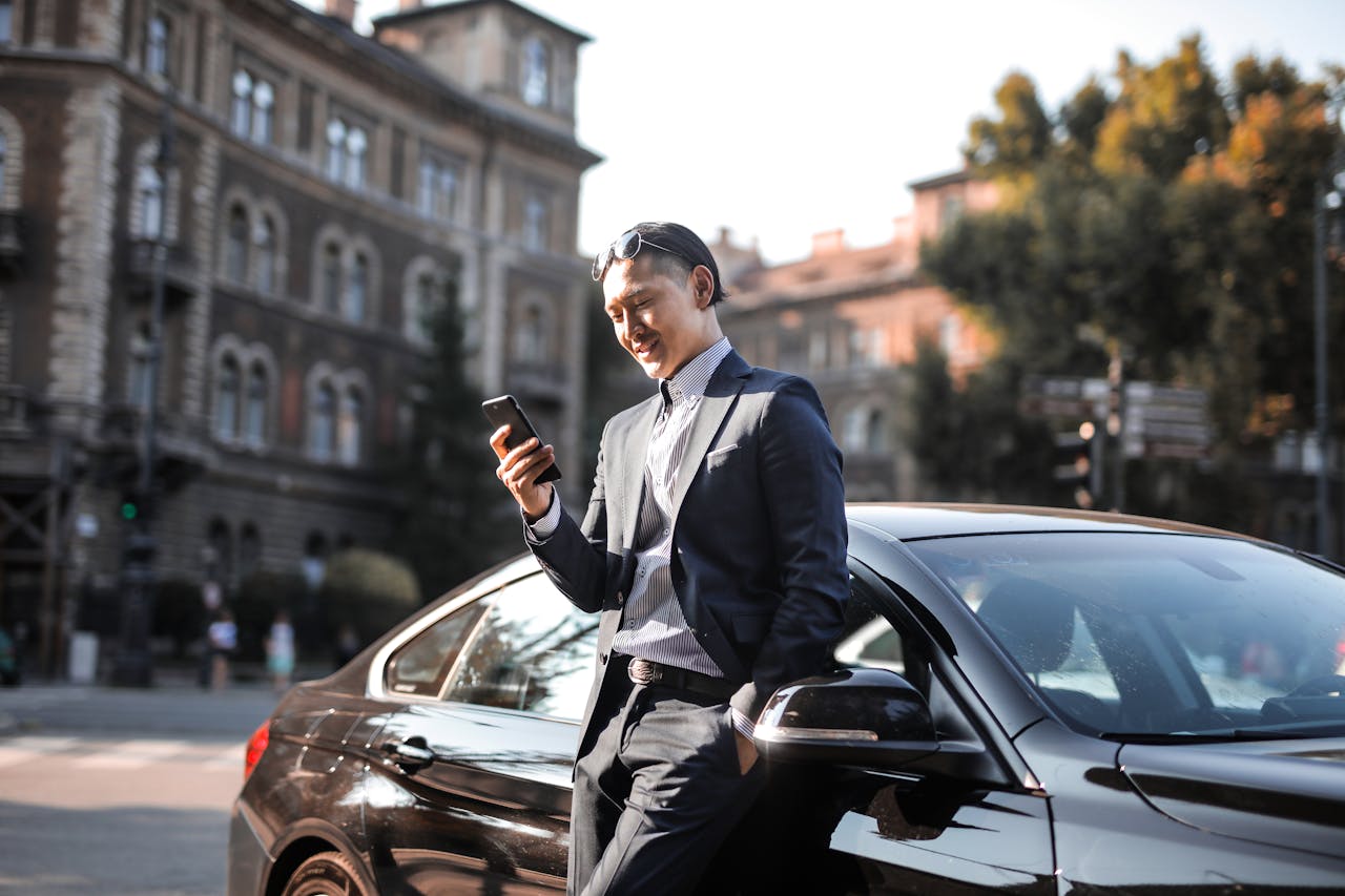  Man in Black Suit Using His Phone While Leaning Beside Black Car