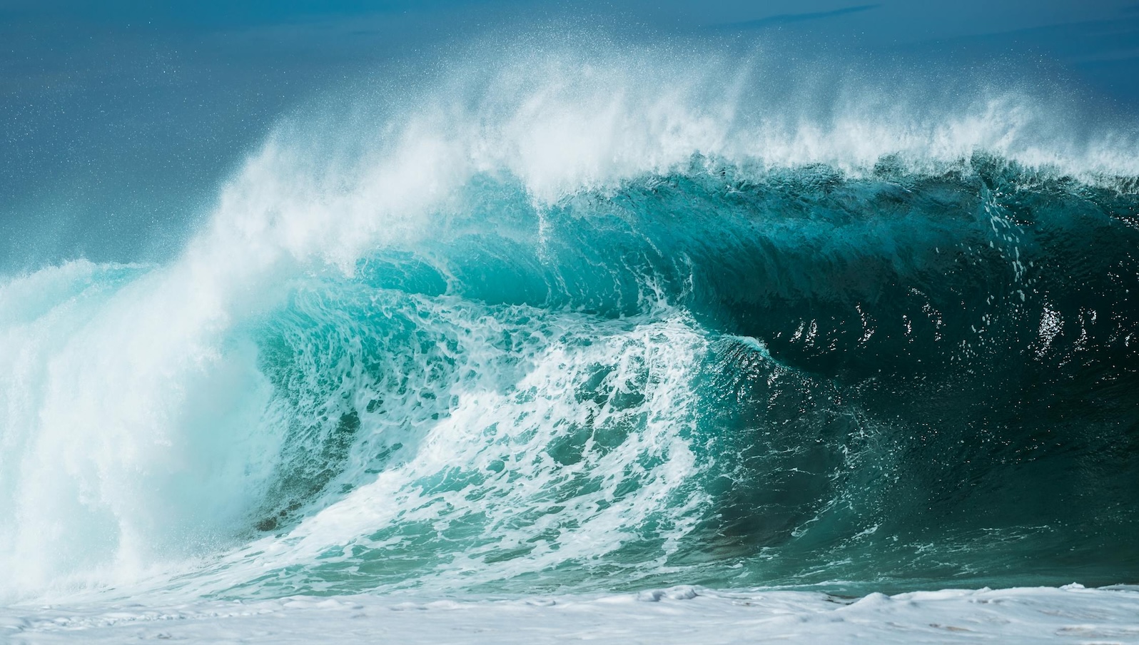 A large teal blue wave starting to crash into the sea against a blue sky background