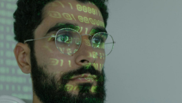 Man with beard and glasses staring off, images of a computer screen reflecting back on his face