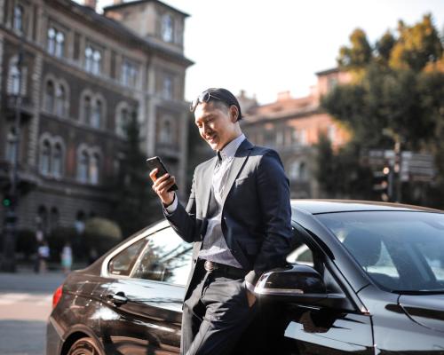  Man in Black Suit Using His Phone While Leaning Beside Black Car